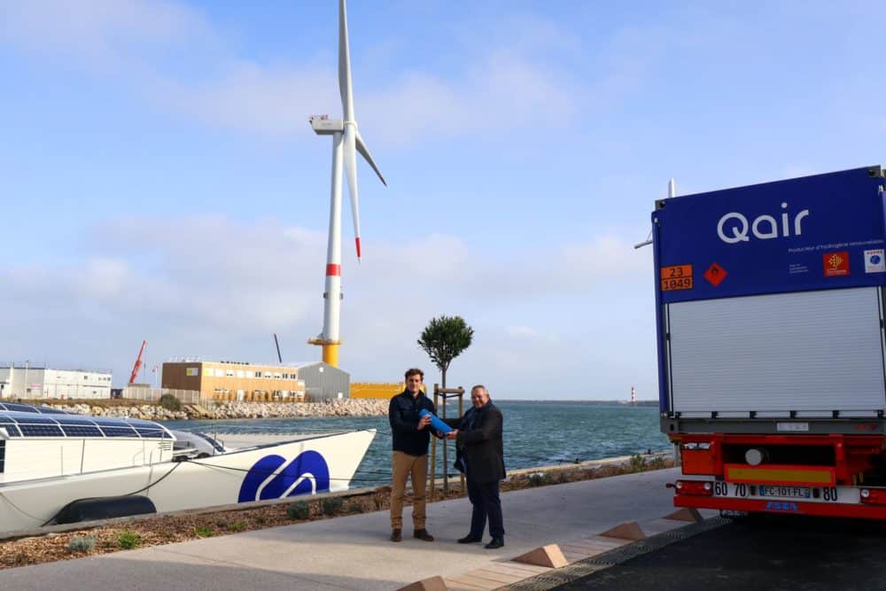 Jean-Sébastien Lasbouygues, Director of the H2 Business Unit at Qair France, symbolically hands over the first molecule produced by Hyd'Occ to Marin Jarry, captain of Energy Observer. The Energy Observer boat and a floating wind turbine from Qair's Eolmed project can be seen in the background.
